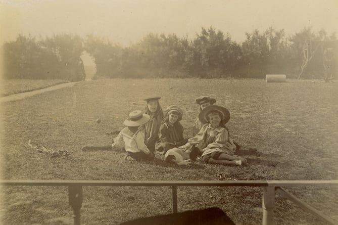 Five children seated on grass all wearing smart clothes and hats. Small dog in centre of group.