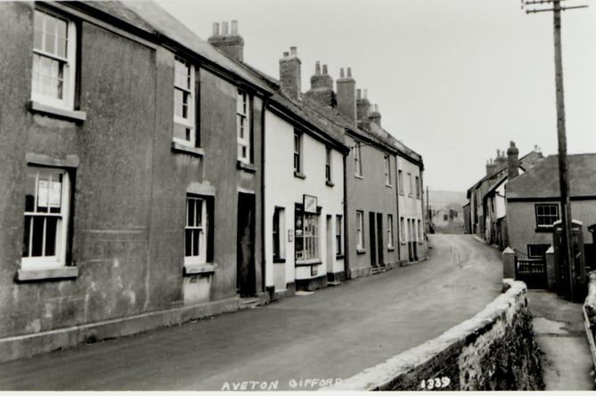 Telephone box on right hand side of Fore Street, Aveton Gifford. Provision store on left. Brick wall in front of telephone box. Around 1910.